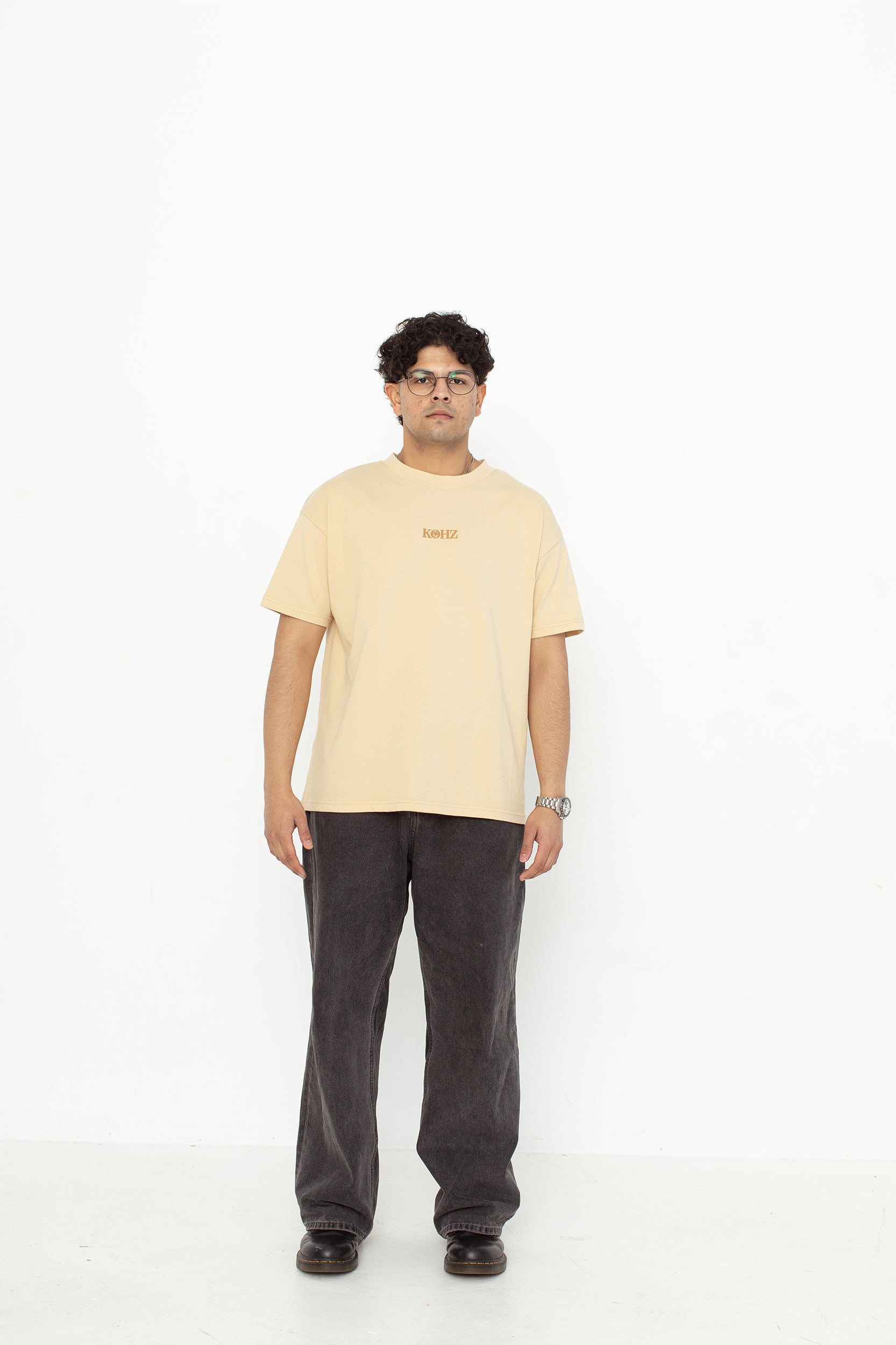 A model wearing a beige oversized kohz bb t-shirt with a puff printed logo, crew neck, and short sleeves standing against a white background.