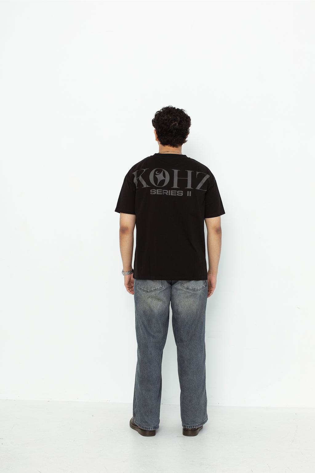 A model wearing a black oversized kohz  t-shirt with a puff printed logo, crew neck, and short sleeves standing against a white background.