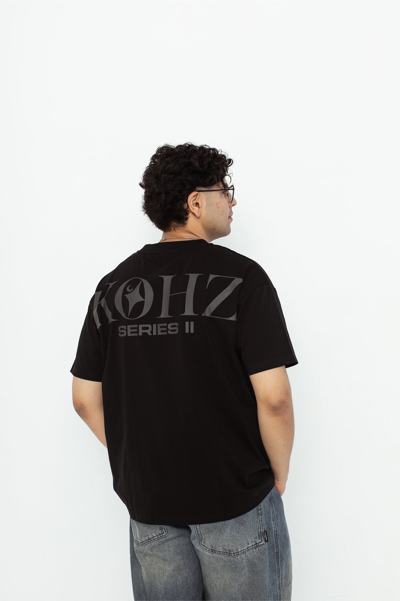 A model wearing a black oversized kohz  t-shirt with a puff printed logo, crew neck, and short sleeves standing against a white background.
