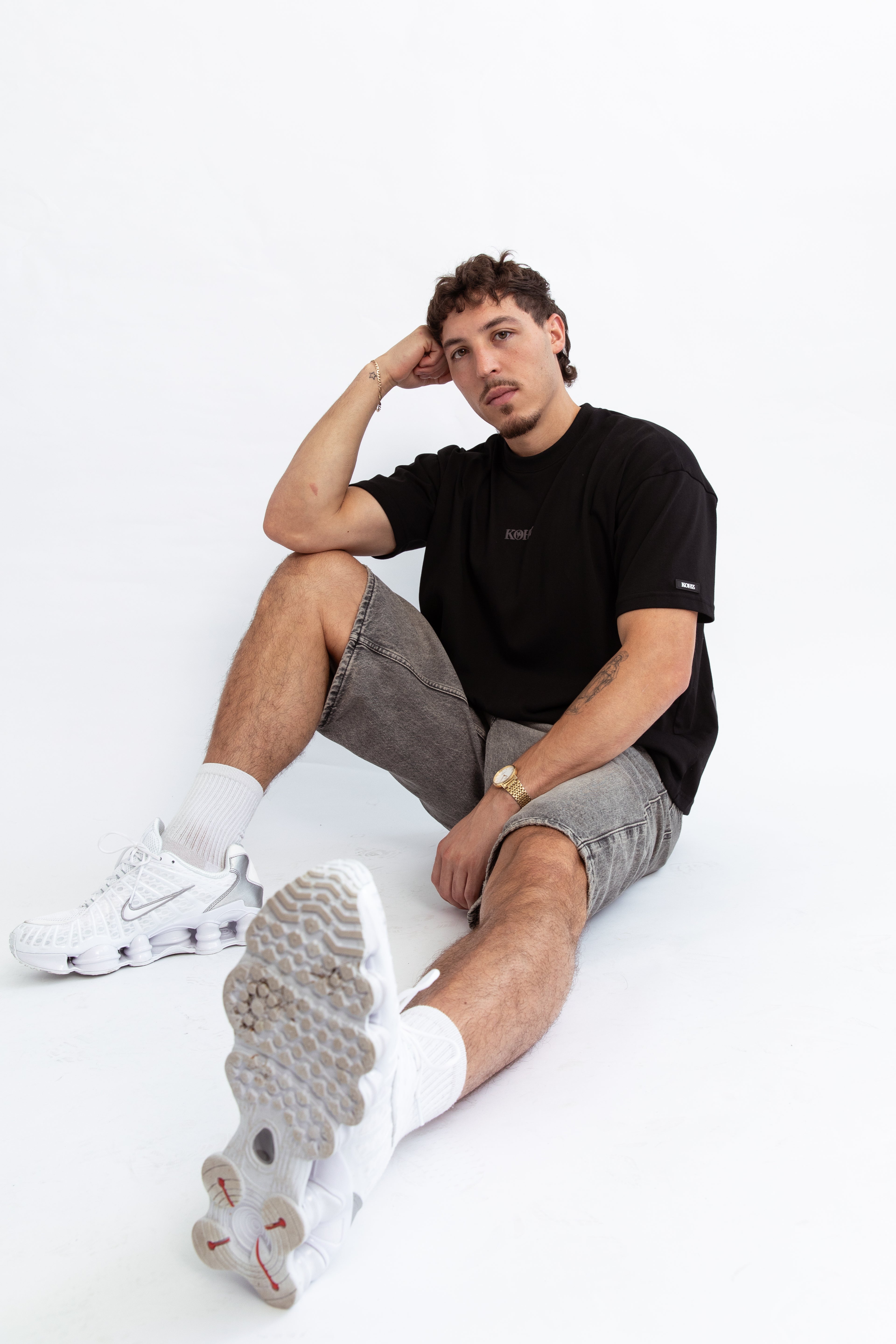 A model wearing a black oversized kohz bb t-shirt with a puff printed logo, crew neck, and short sleeves standing against a white background.
